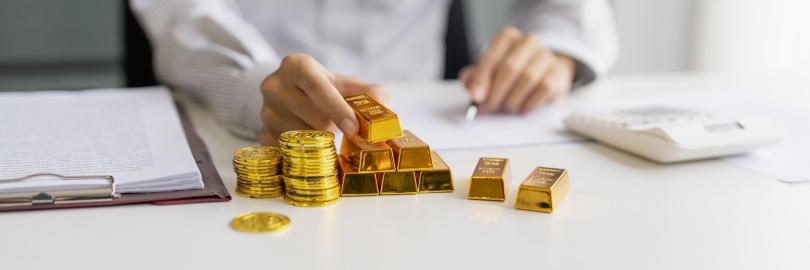 Auf einem Schreibtisch liegen Goldbarren und Münzen, jemand sitz am Schreibtisch mit Taschenrechner
