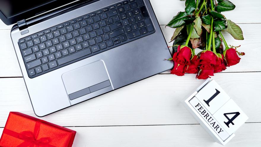 Neben einem Laptop liegen roten Rosen und ein verpacktes Geschenk auf einem Tisch.