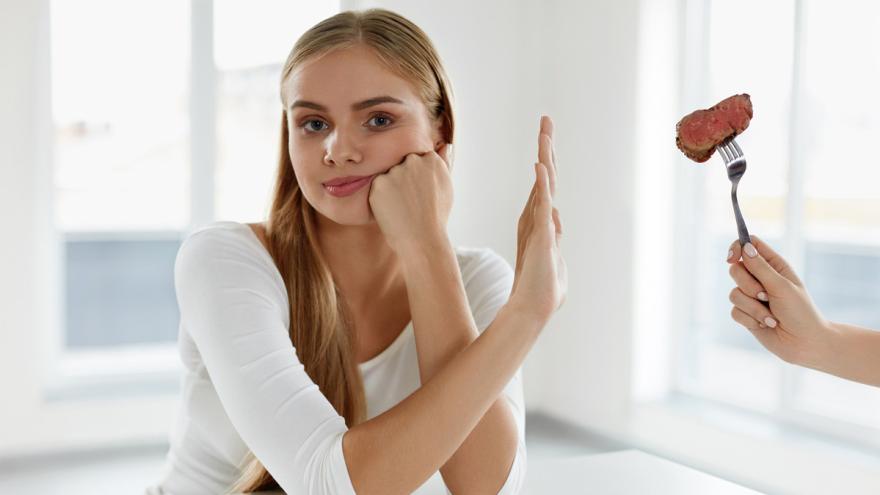 Frau hält Hand ablehnend vor Gabel, die ihr mit einem Stück Fleisch von der Seite gereicht wird