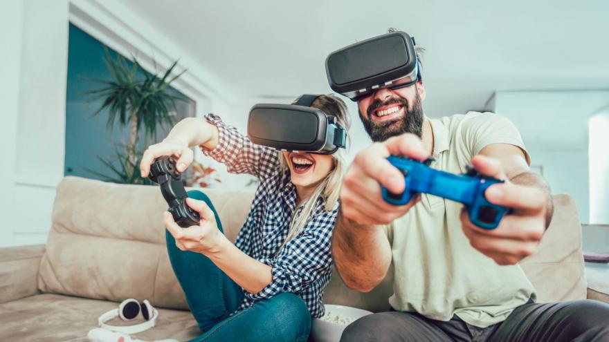 Ein junges Paar sitzt lachend auf einem beigen Sofa und trägt Virtual-Reality-Headsets. Beide halten Gaming-Controller in den Händen und scheinen mitten in einem Videospiel zu sein. Die Frau trägt ein kariertes Hemd und gepunktete Socken, während der Mann ein helles Poloshirt trägt. Vor ihnen auf dem Sofa liegen Kopfhörer und eine Schüssel Popcorn.