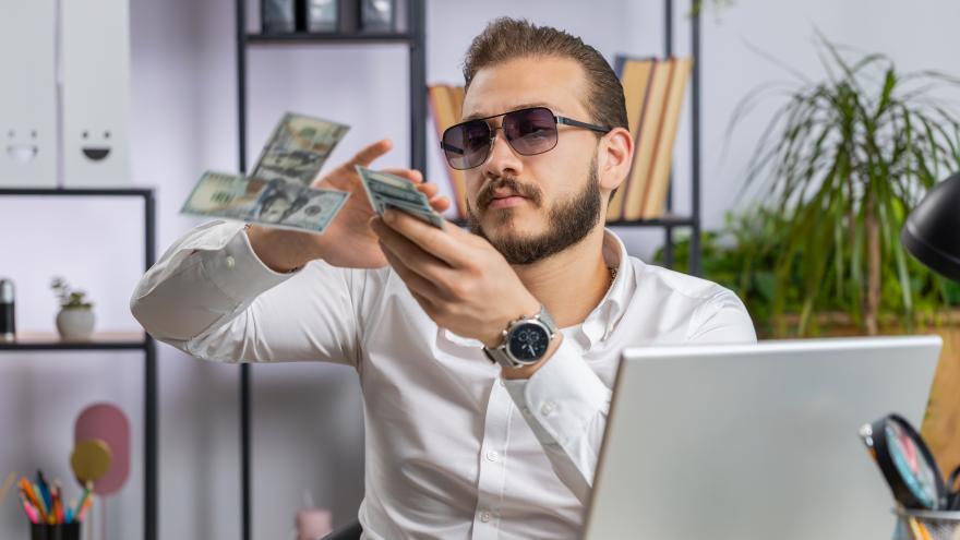 Ein Mann mit Sonnenbrille sitzt vor einem Laptop und schmeißt mit Geldscheinen.