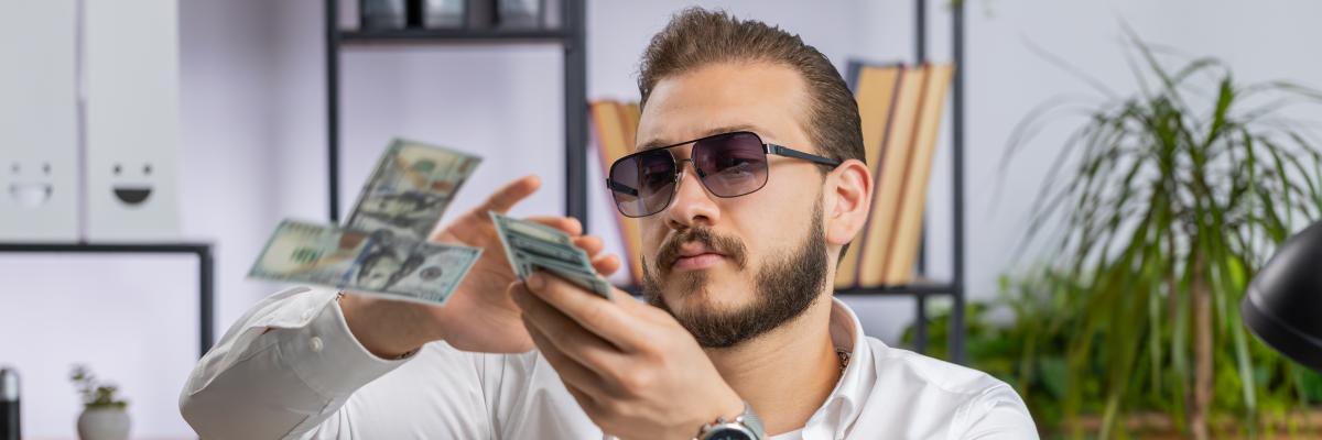 Ein Mann mit Sonnenbrille sitzt vor einem Laptop und schmeißt mit Geldscheinen.