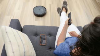 Blick von leicht oben über die Schulter einer Person, die auf einem Sofa sitzt und eine Fernbedienung nutzt. Neben ihr liegen ein Smartphone und ein Kissen, auf dem Boden befindet sich ein Staubsaugerroboter