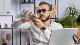 Ein Mann mit Sonnenbrille sitzt vor einem Laptop und schmeißt mit Geldscheinen.